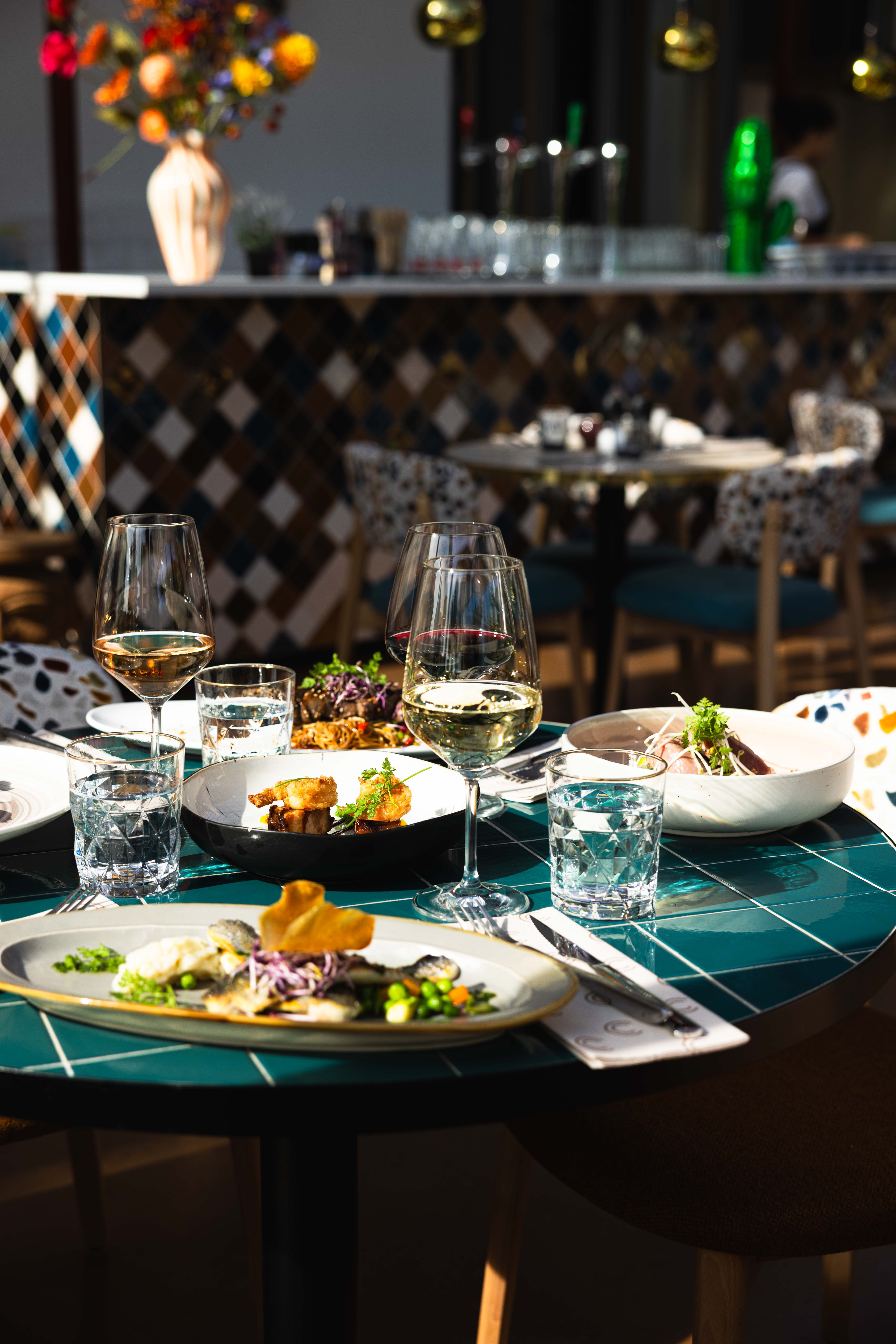 Gedekte tafel bij La Cocotte met gerechten om te delen, gegrilde zeebaars, een glas wijn en de mozaïekbar op de achtergrond
