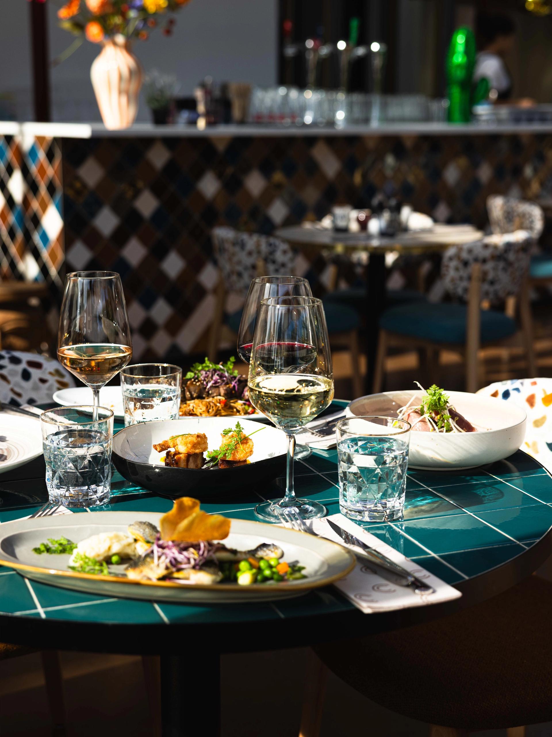 Gedekte tafel bij La Cocotte met gerechten om te delen, gegrilde zeebaars, een glas wijn en de mozaïekbar op de achtergrond
