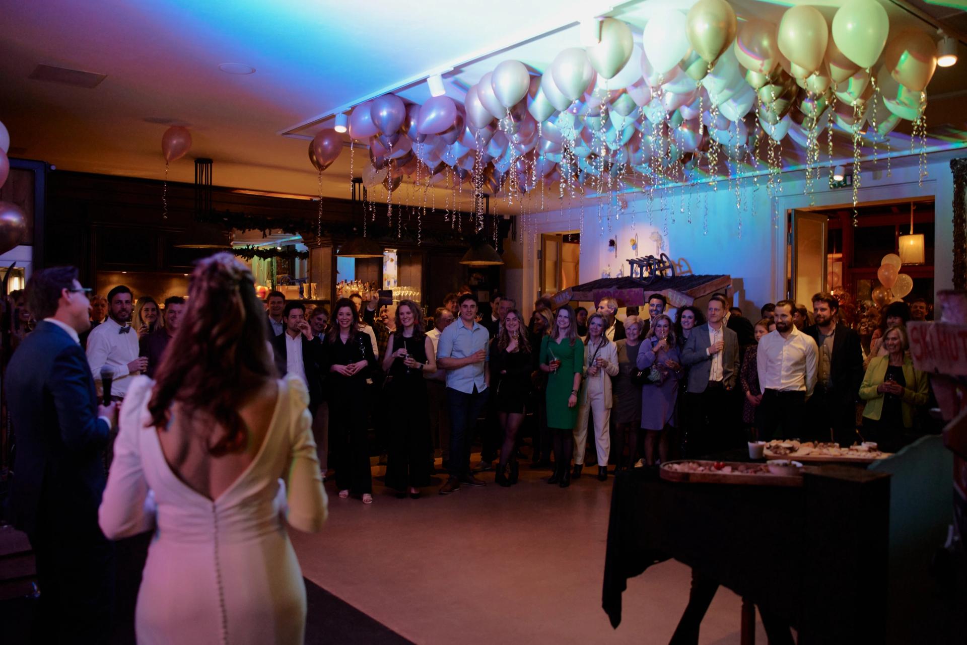 Bruidspaar wordt toegesproken op het feest in het restaurant, met gasten in een halve kring onder een plafond vol ballonnen