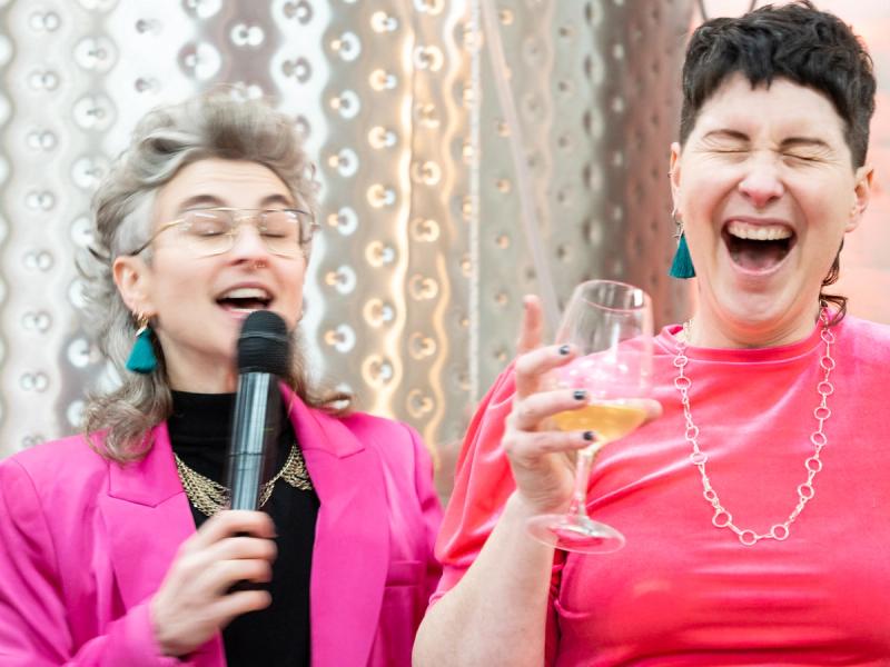 two people wearing pink laughing. one person is speaking into a microphone