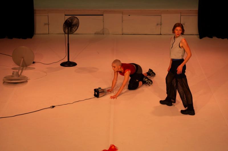 one dancer on a stage stands and looks behind them, while another dancer is on all fours with one hand on a blender