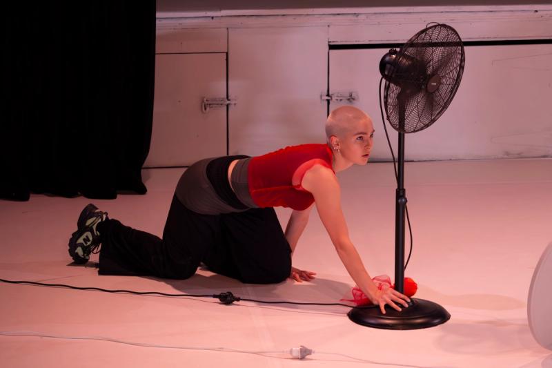 a dancer is on all fours on a white stage. they are wearing a red top, black pants, runners and have a shaved blond head. they have one hand on a black fan in front of them