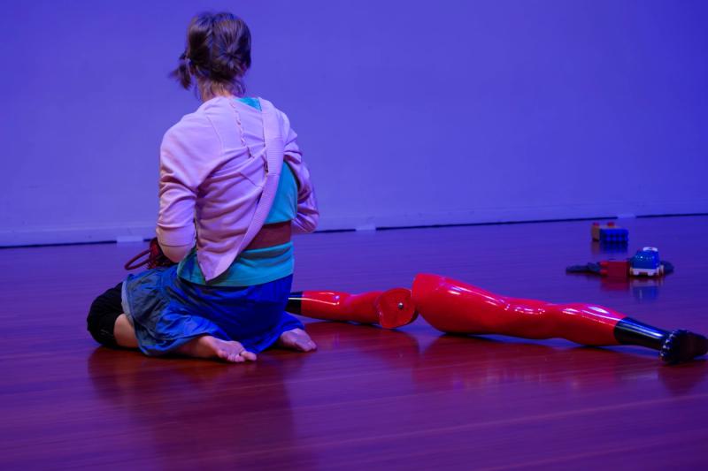 a dancer in purple lighting sits with their back to the camera, looking at toys next them
