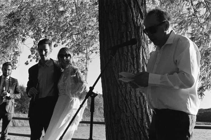a bride and groom listen to an older man making a speech at a wedding on a lake