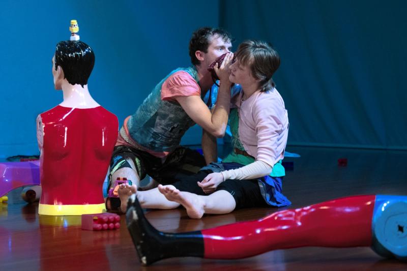 two dancer sit on stage with toys surrounding them. one whispers into the other one's ear