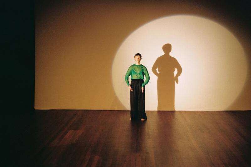 a person wearing a green top and black pants against a white backdrop stands in a spotlight