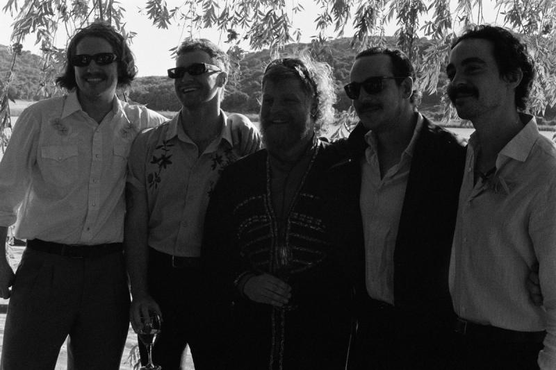 five men in suits and sunglasses smile with their arms around each other in front of a lake