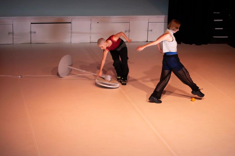 two dancers are on a stage. one dancer leans over holding a fan, another dancer jumps over an orange