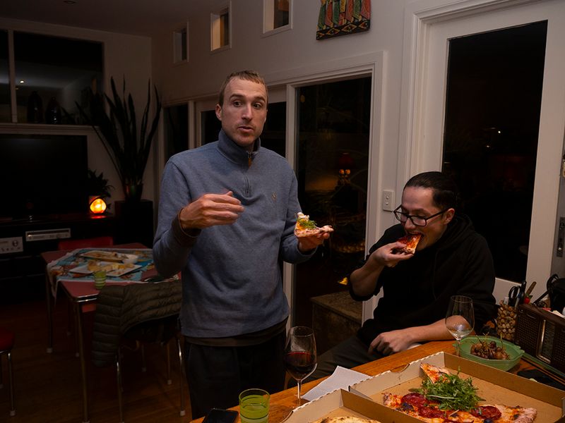 two men at a kitchen bench eating pizza