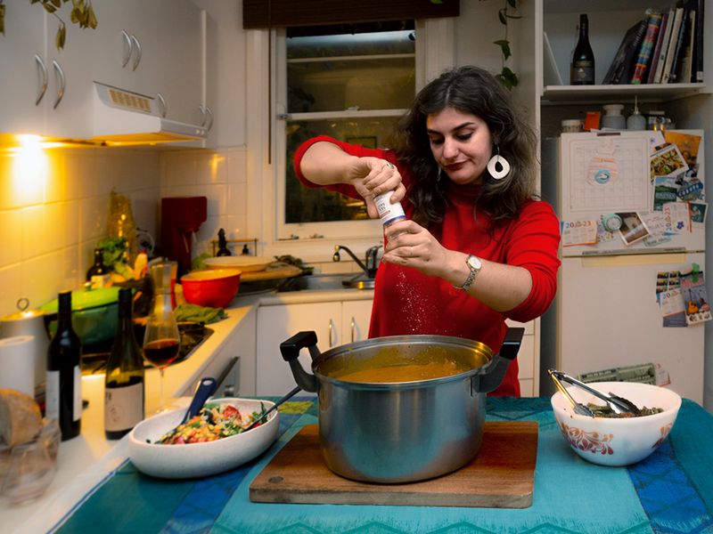 a portrait of a woman in a kitchen putting salt in a pot of soup