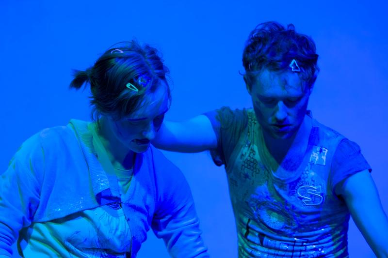 two performers in blue lighting sit next to each other. one has their hand on the other one's neck and both are looking down