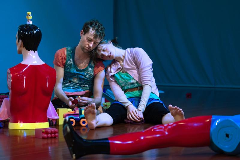 two performers sit on the floor surrounded by toys. one has their legs spread in front them and their head on the other person's shoulder