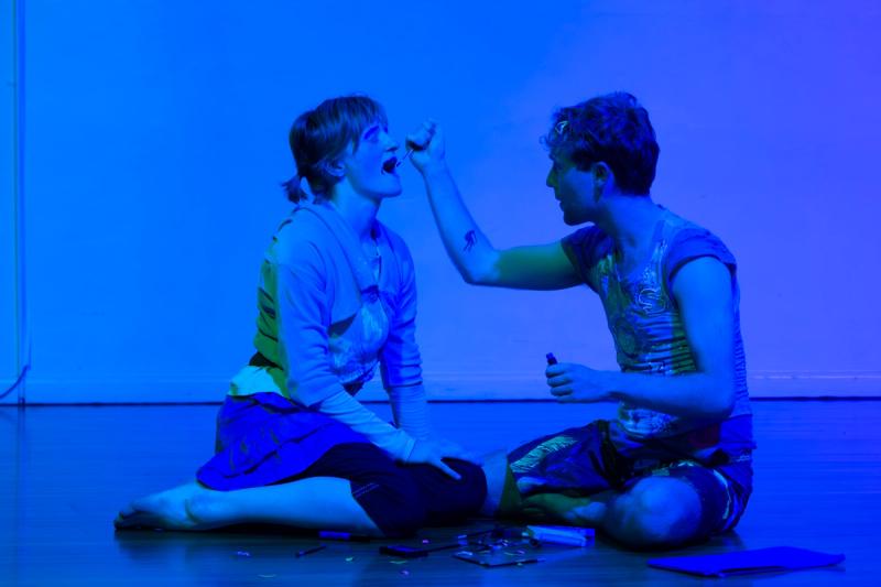two performers sit on a floor in blue lighting. one is putting lipstick on the other performer's lips