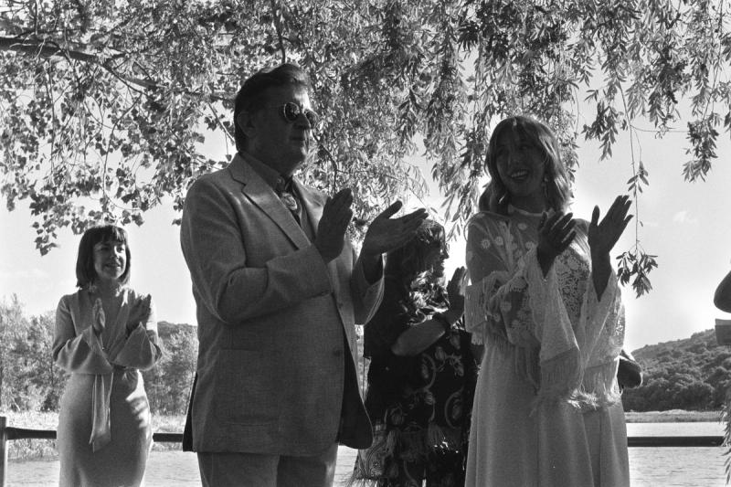 a bride smiles at her father dressed in a suit as they clap next to each other under a tree