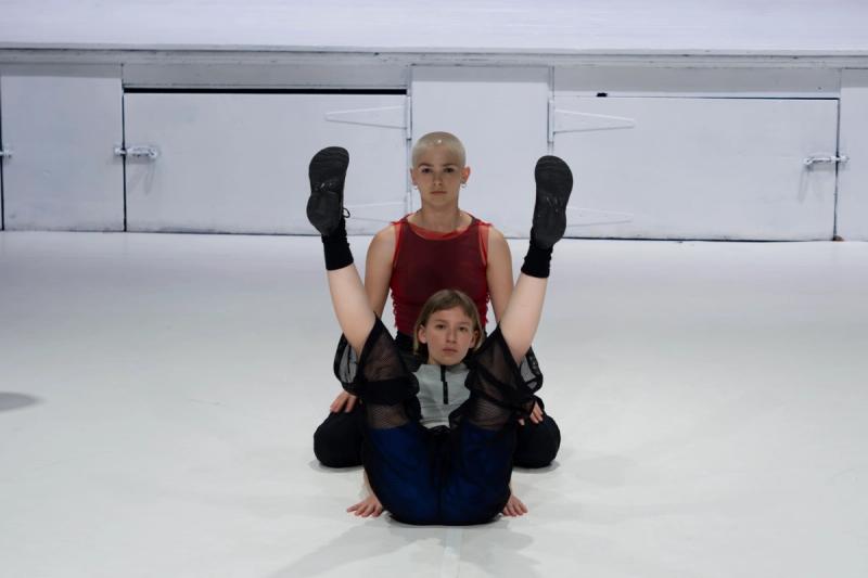 two dancers on a white stage face and look into the camera. one sits behind the other with their knees spread wide and their hands on their knees. the other sits in front of them with their legs in the air