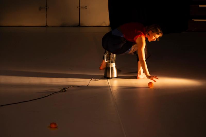 a performer on stage sits on a blender with a side spotlight on them. One of their hands reaches for an orange on the floor behind them