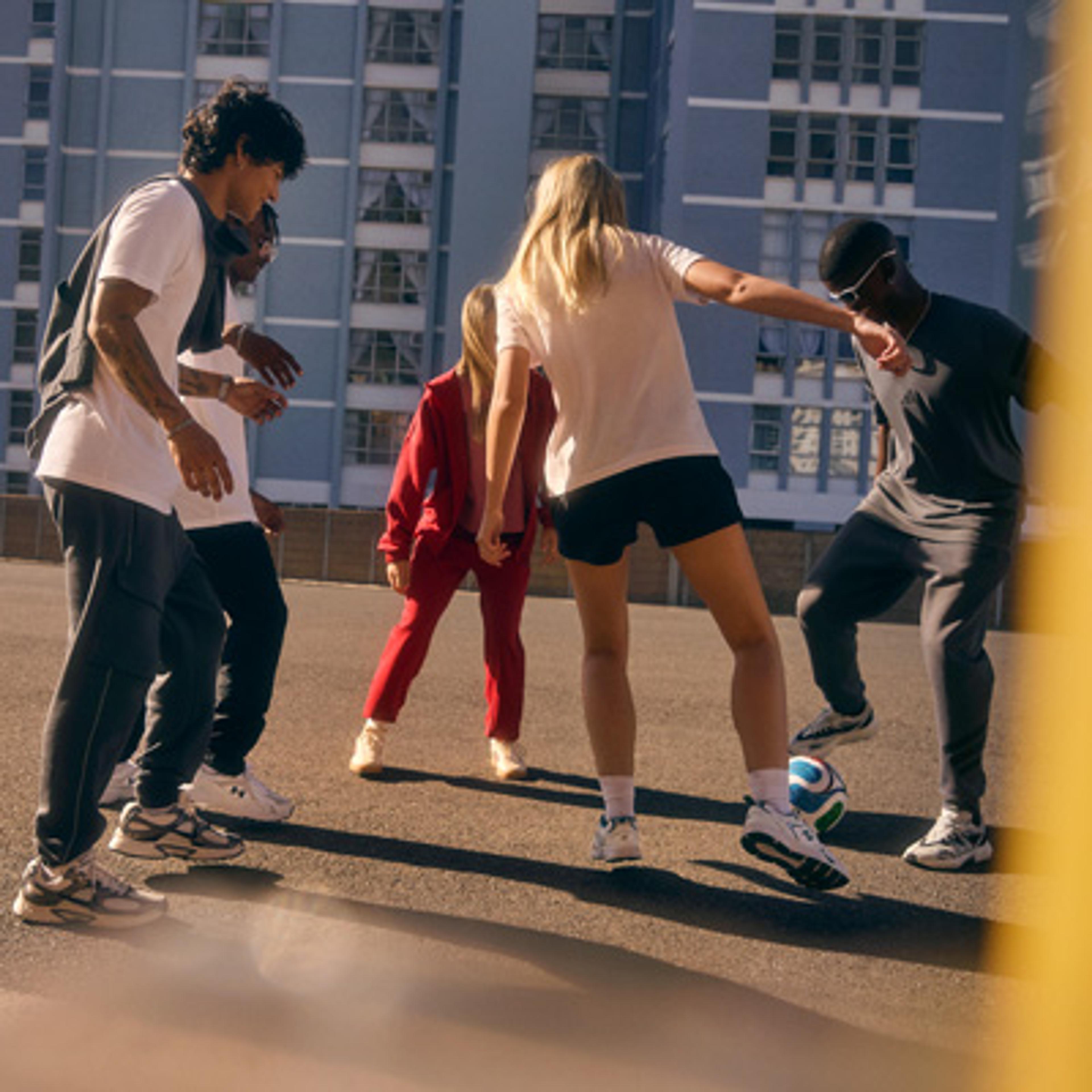 Five people playing soccer on an outdoor asphalt court with a tall blue building in the background.