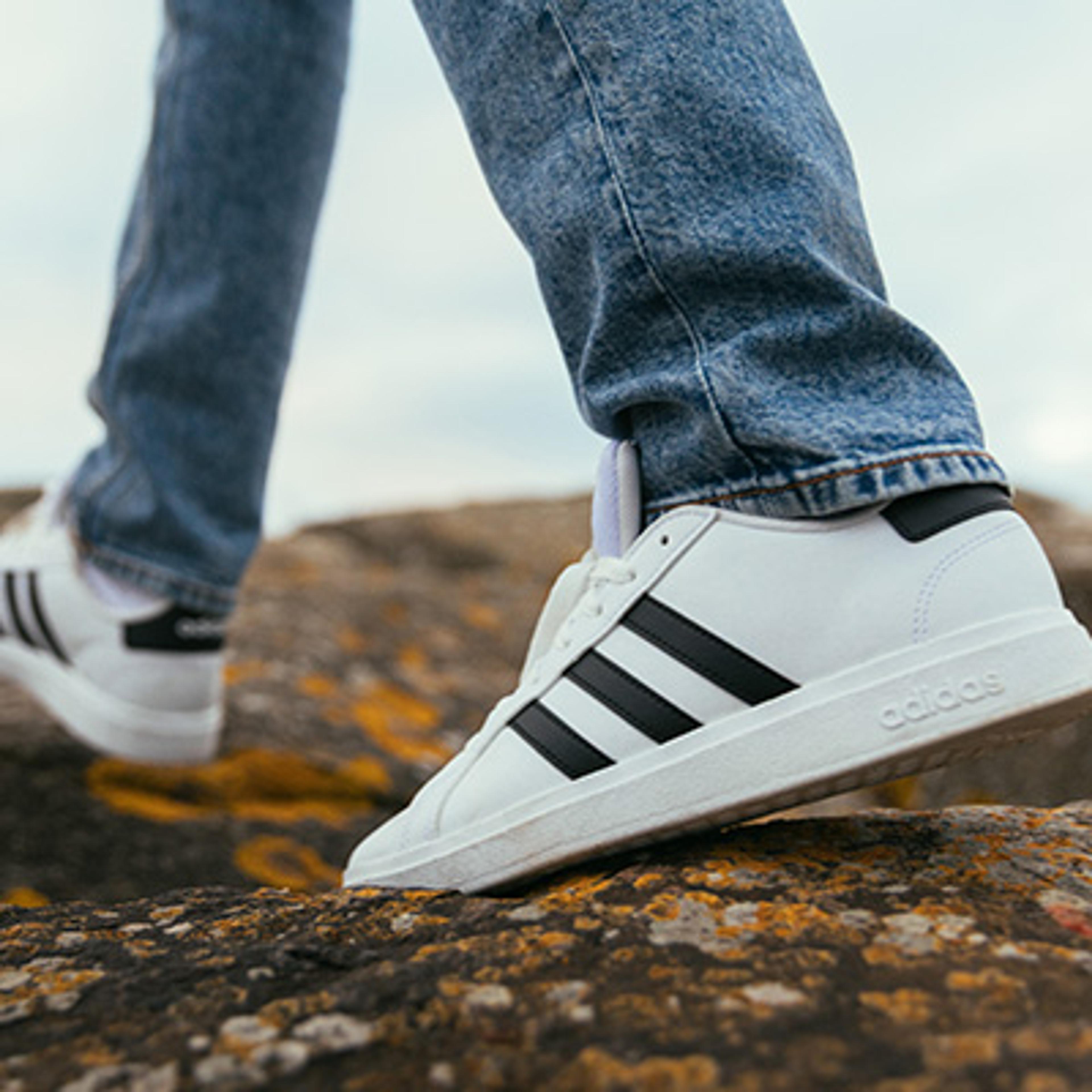 Person in blue jeans and white Adidas sneakers walking on rocky ground.