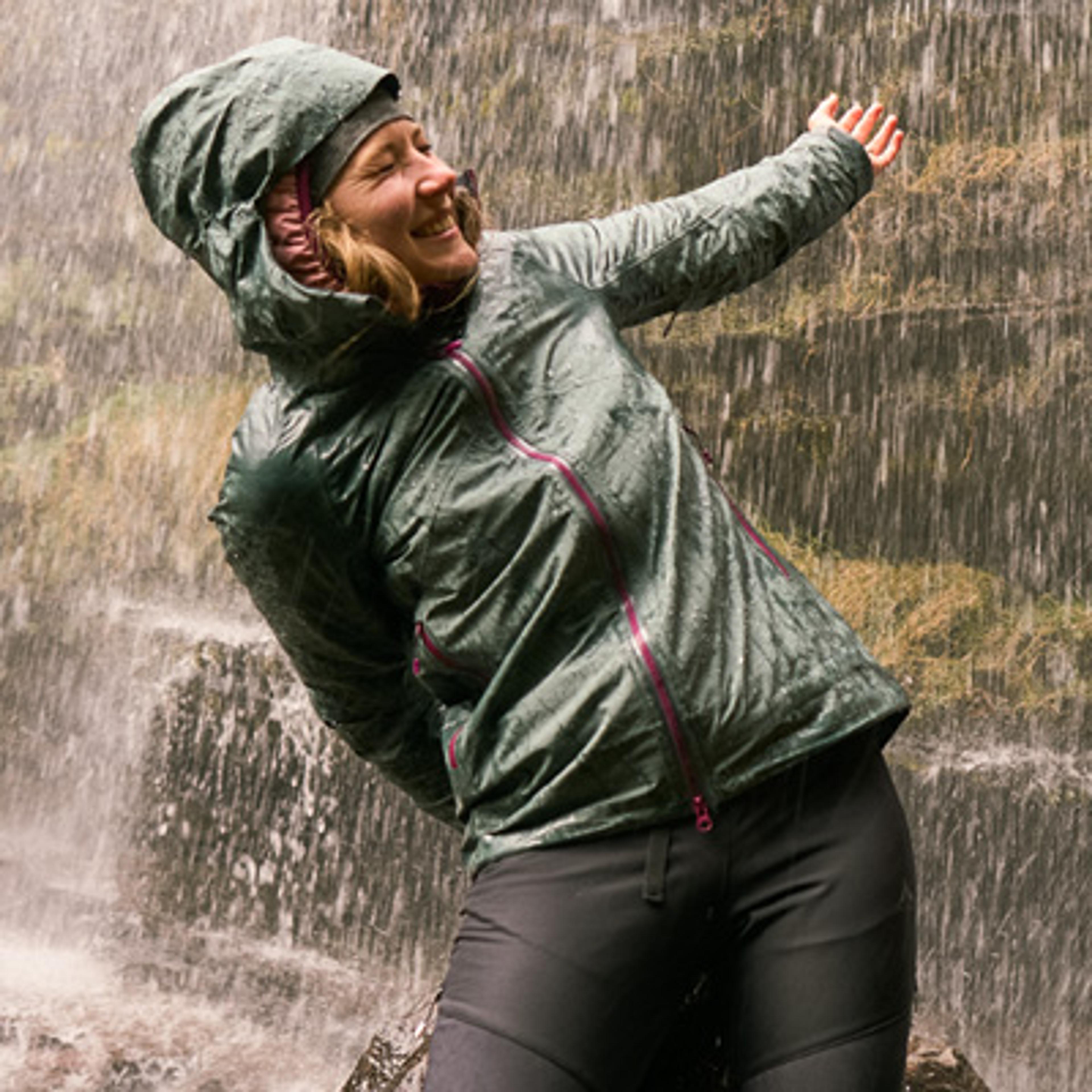 Smiling woman in a dark green hooded rain jacket reaching out under a waterfall.