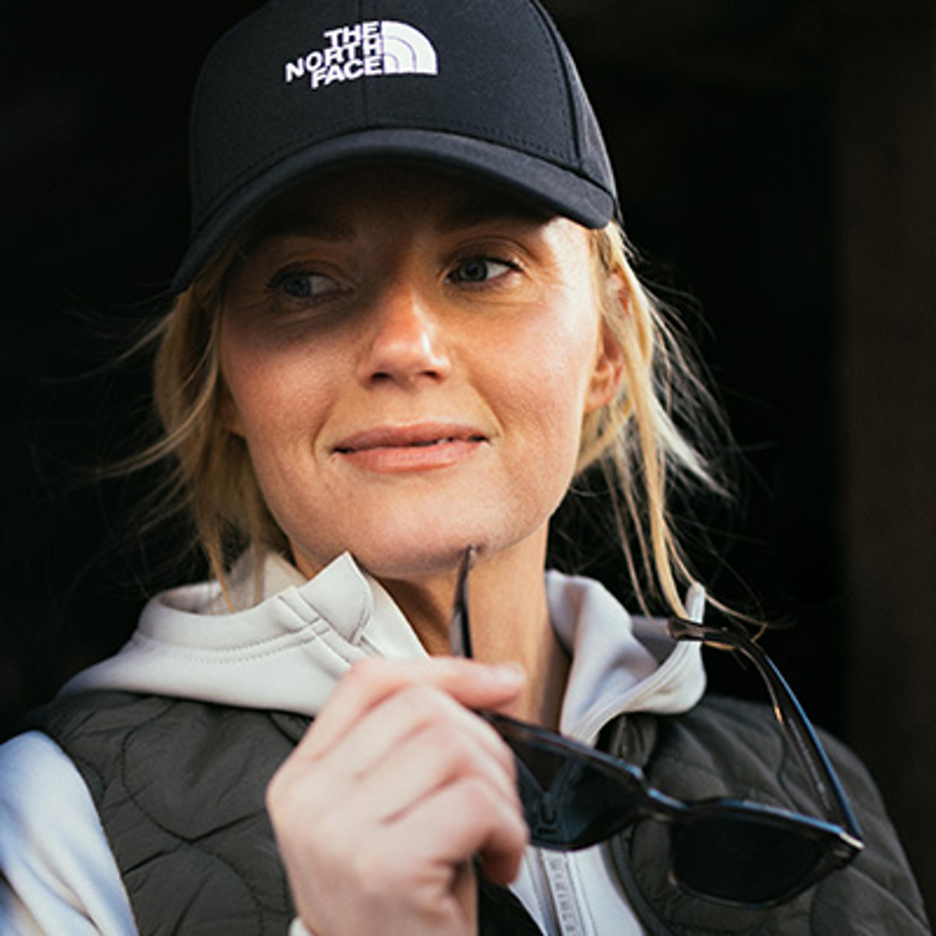 A woman in a North Face cap and quilted vest holds sunglasses, looking sideways with a slight smile.