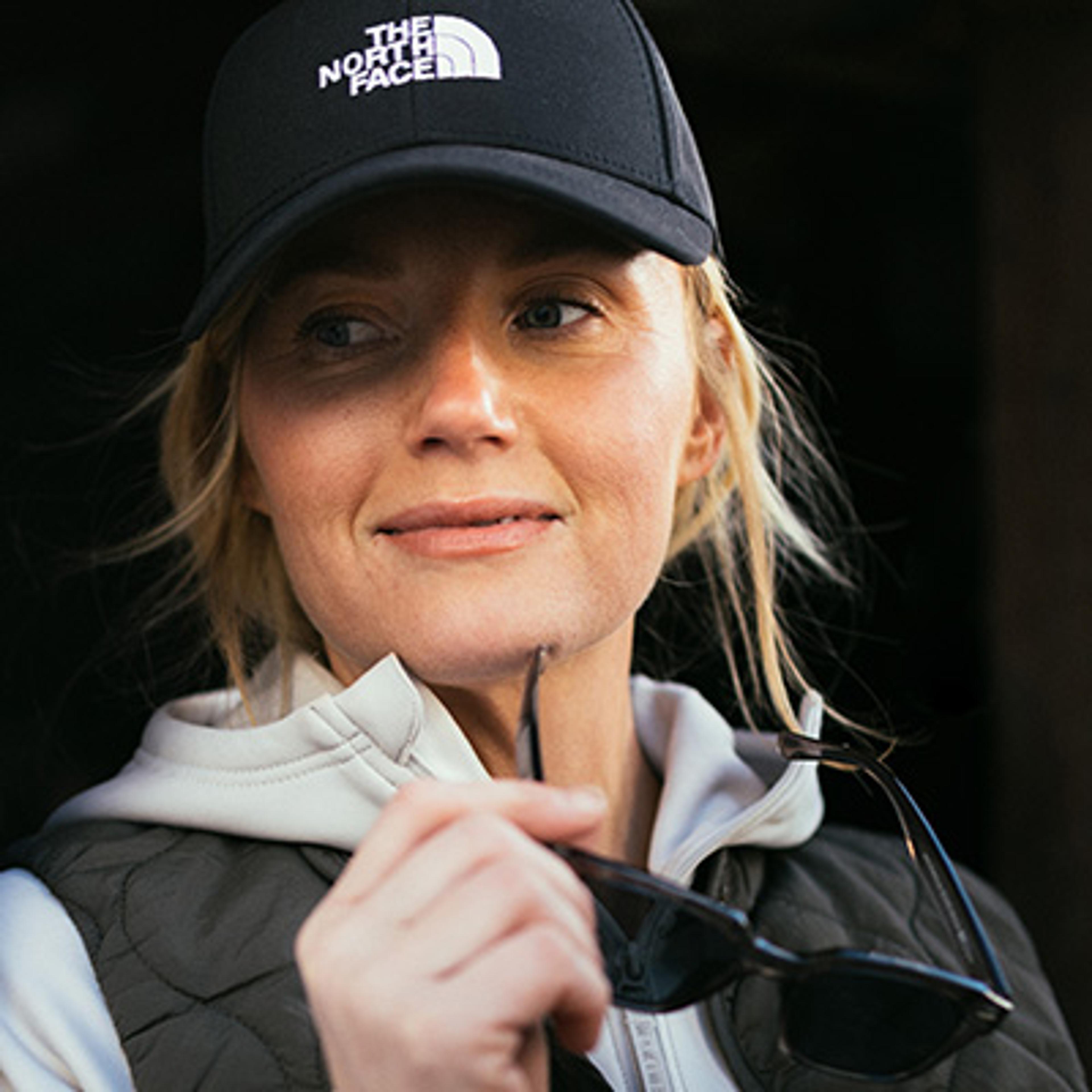 A woman in a black The North Face cap holds sunglasses and smiles, looking away.