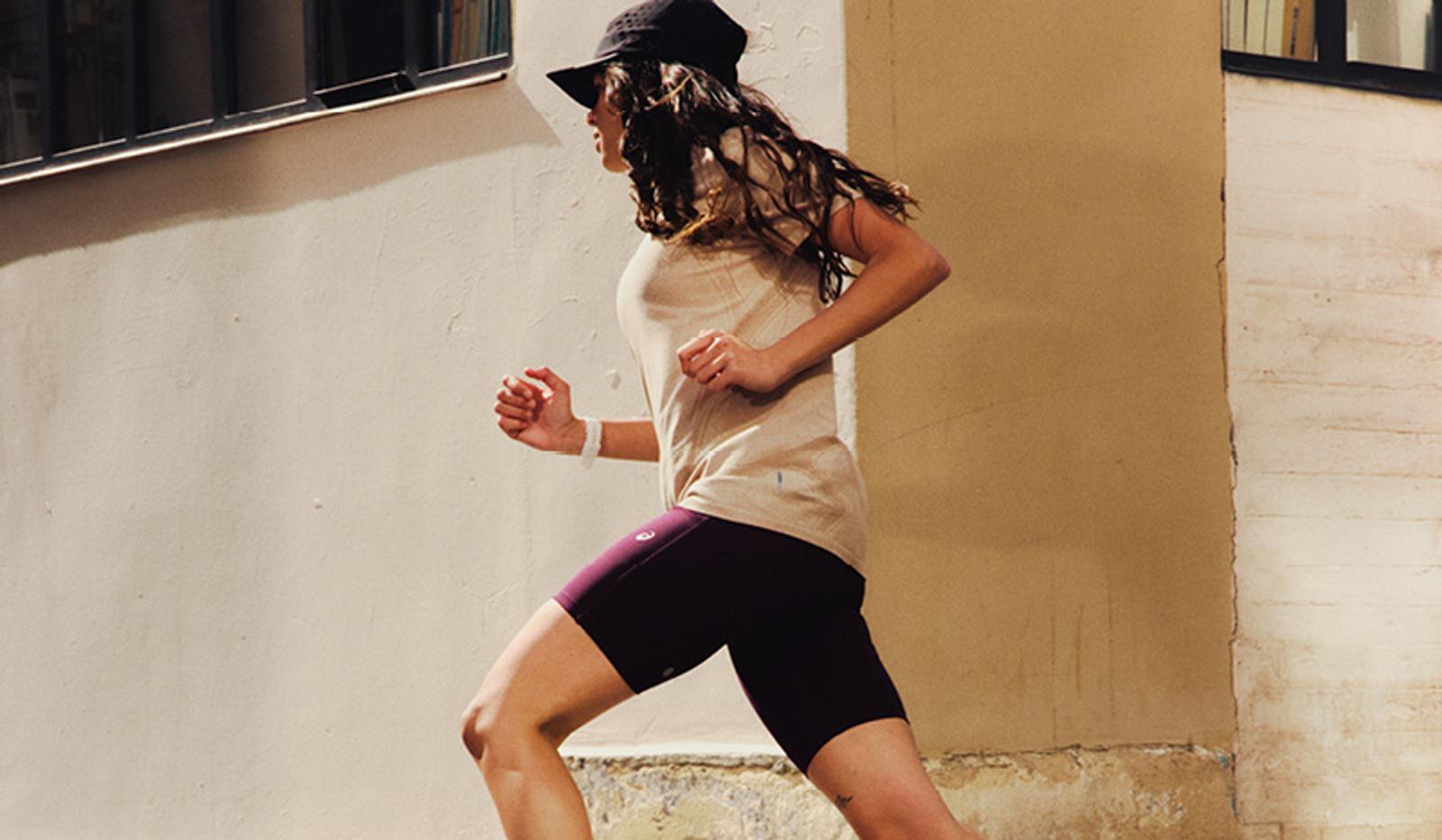 A woman in a black cap, light t-shirt, and dark shorts runs past a light-colored wall.
