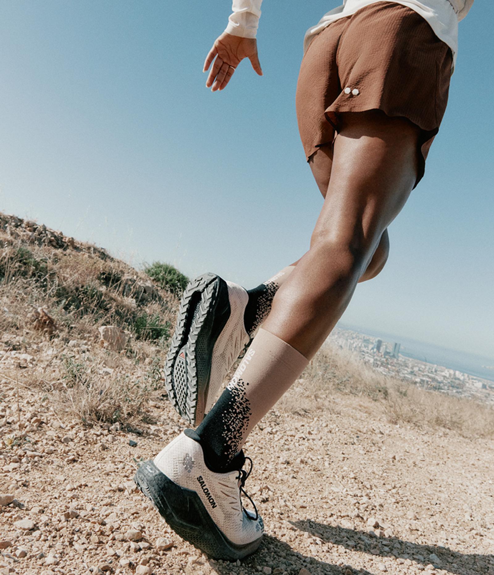 Legs of a runner in motion on a dusty trail, wearing Salomon shoes and socks, with a distant city and blue sky in the background.