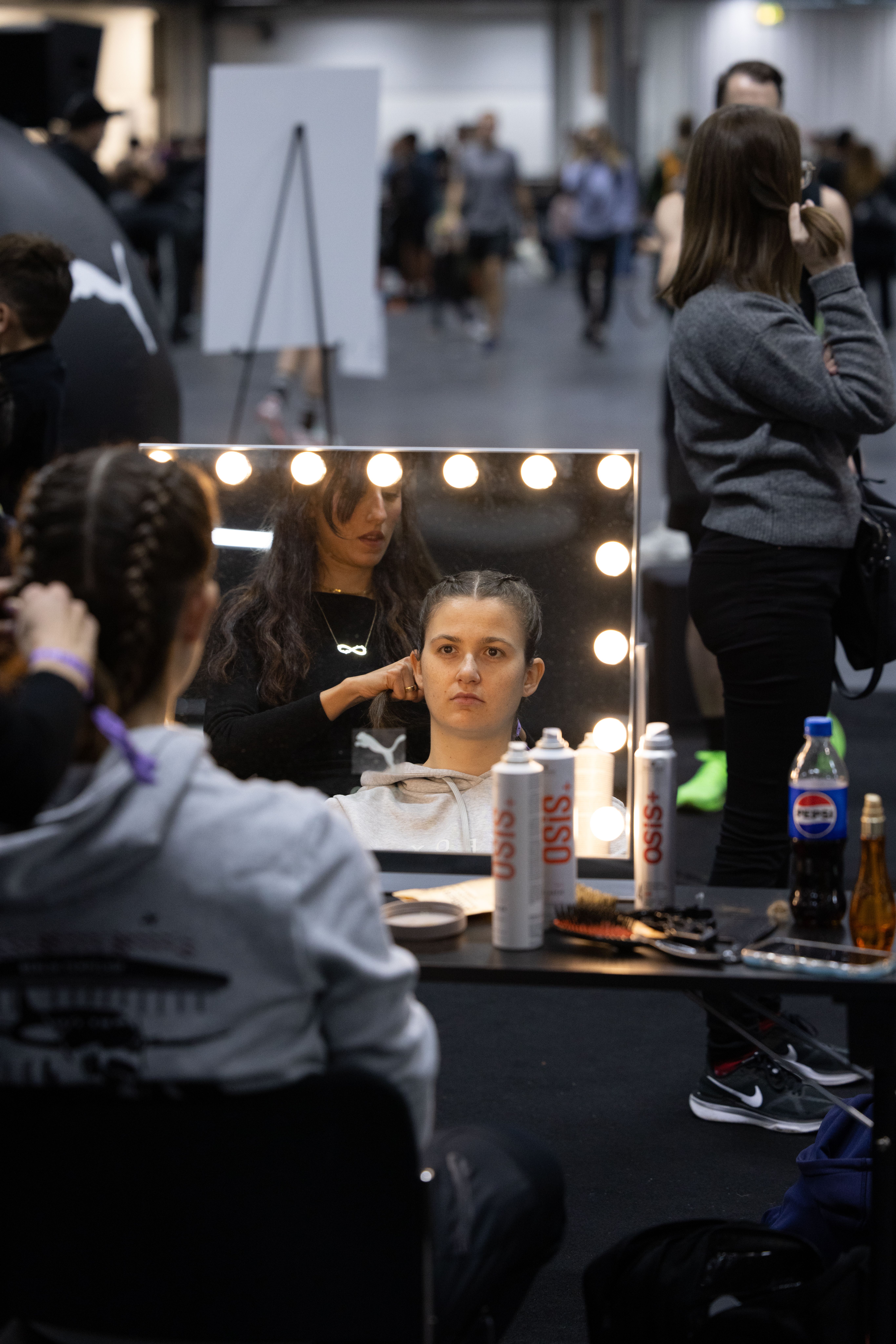 A stylist braids a woman's hair, reflected in a lighted mirror with hair products on a table, in a blurry event space.