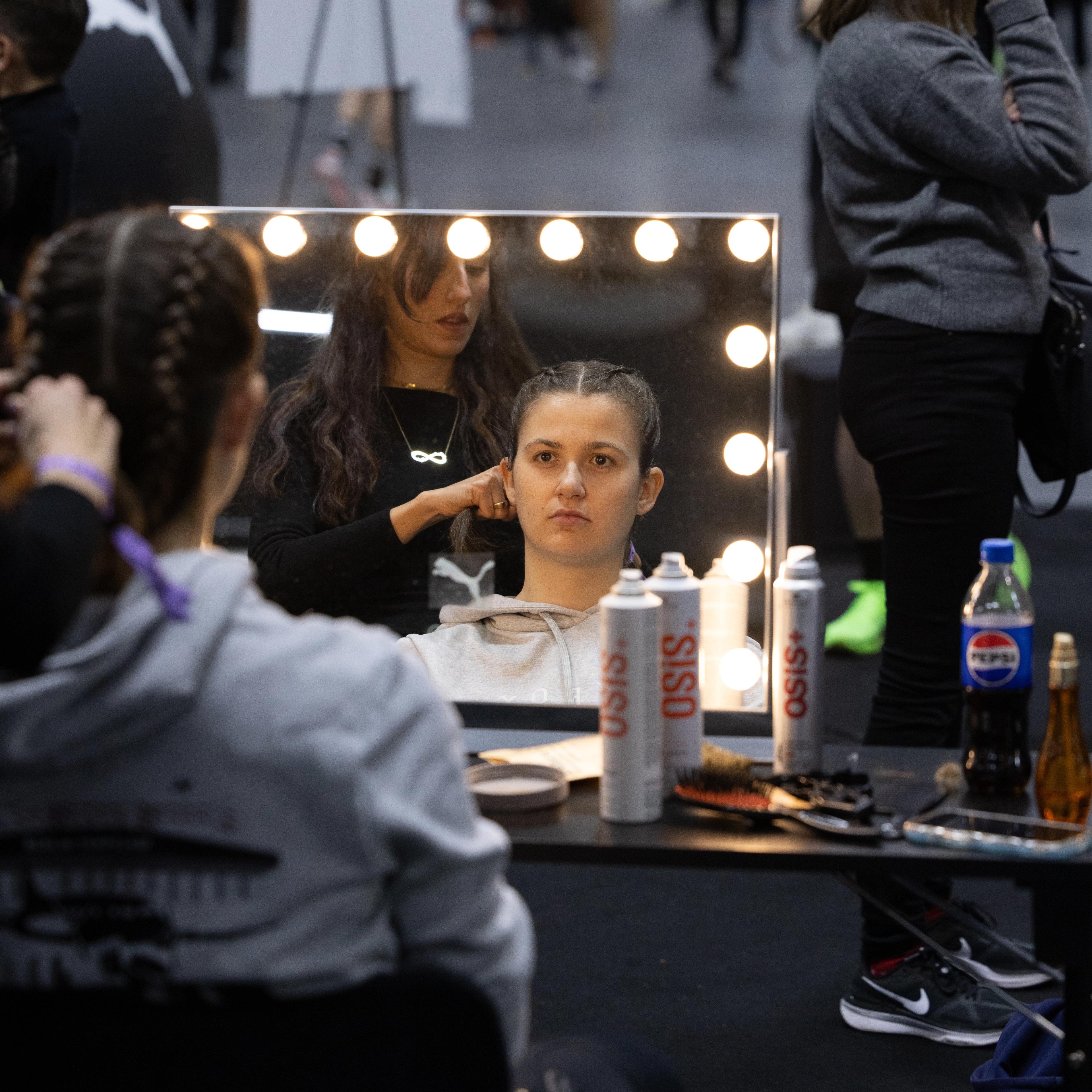 A stylist braids a woman's hair, reflected in a lighted mirror with hair products on a table, in a blurry event space.