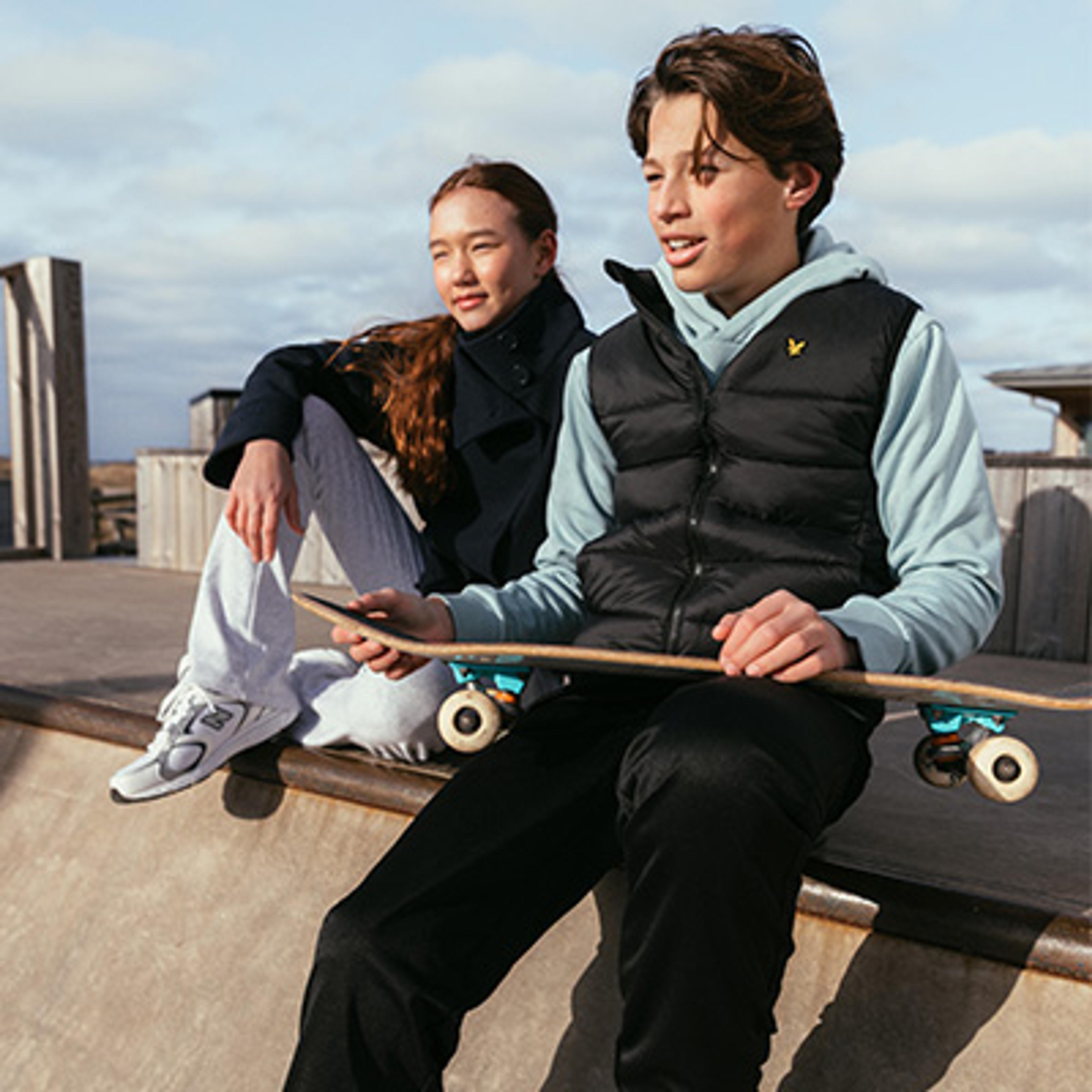 Two teenagers sitting at a skatepark, a boy holding a skateboard.