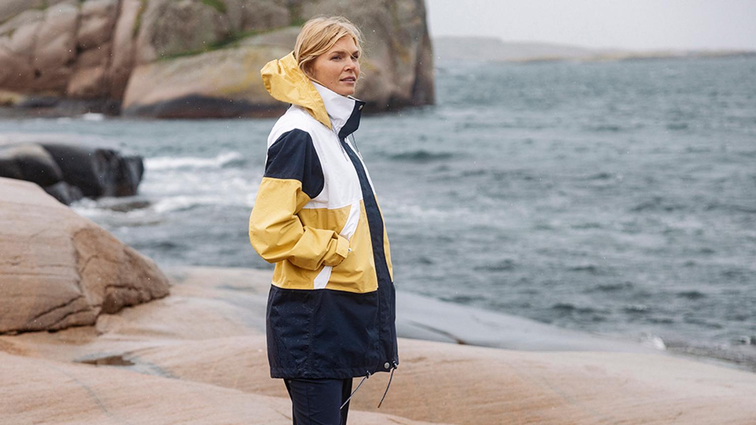 a woman in a yellow and black jacket is standing on a rock near the ocean .