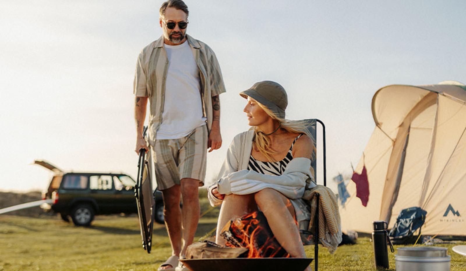 A man stands holding a chair while a woman sits by a campfire at a sunny campsite with a tent and car.