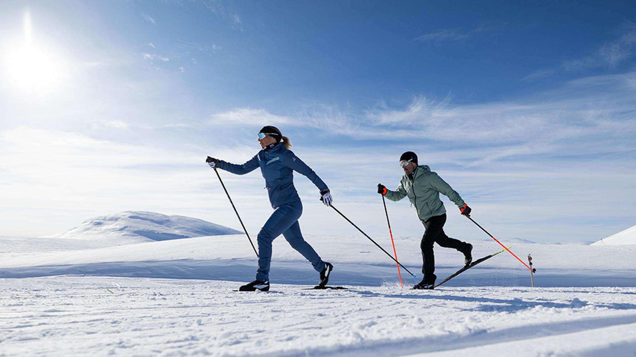 Two people cross-country skiing across a vast snowy landscape on a sunny day.