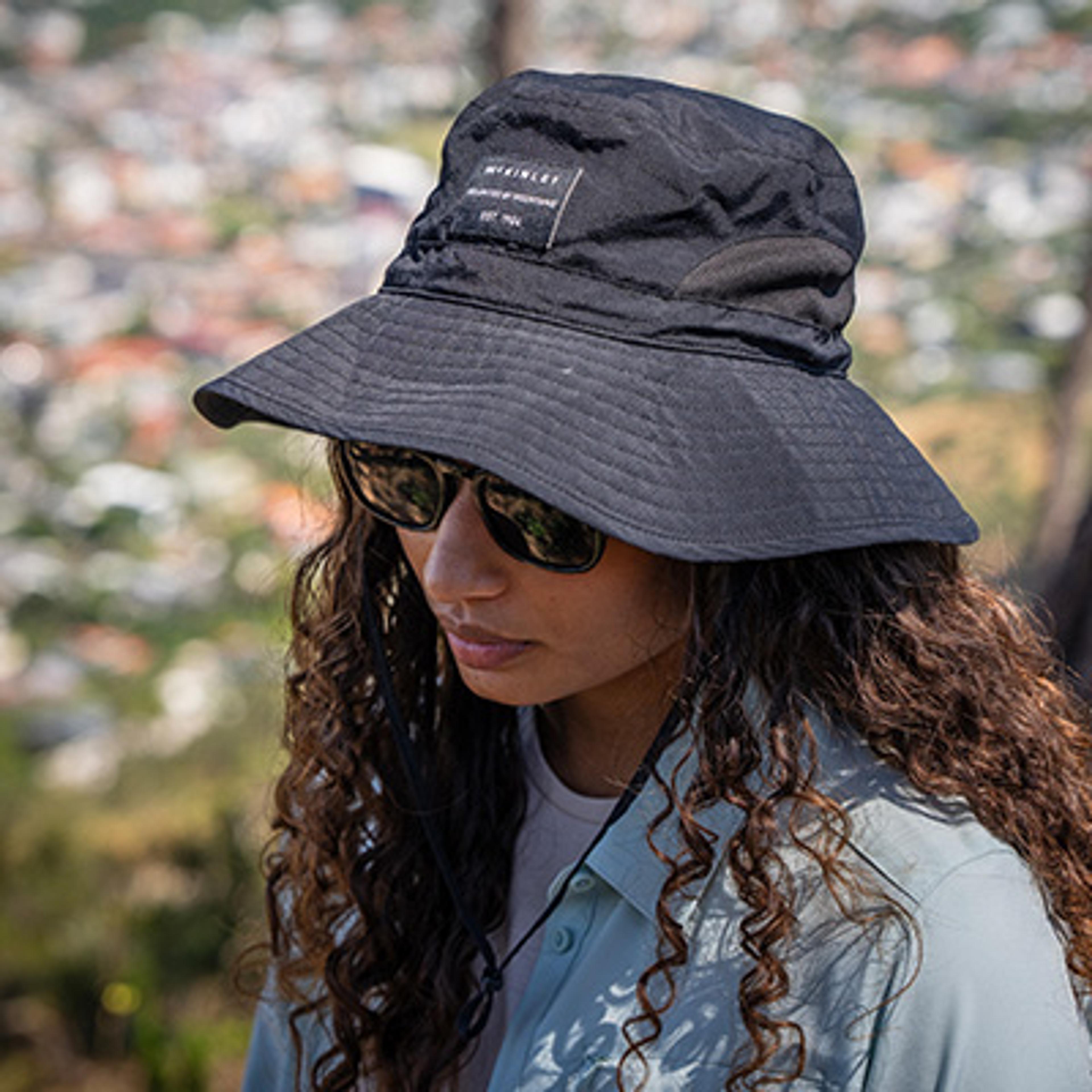 A woman with long curly hair wearing a black McKinley bucket hat, sunglasses, and a light blue shirt.
