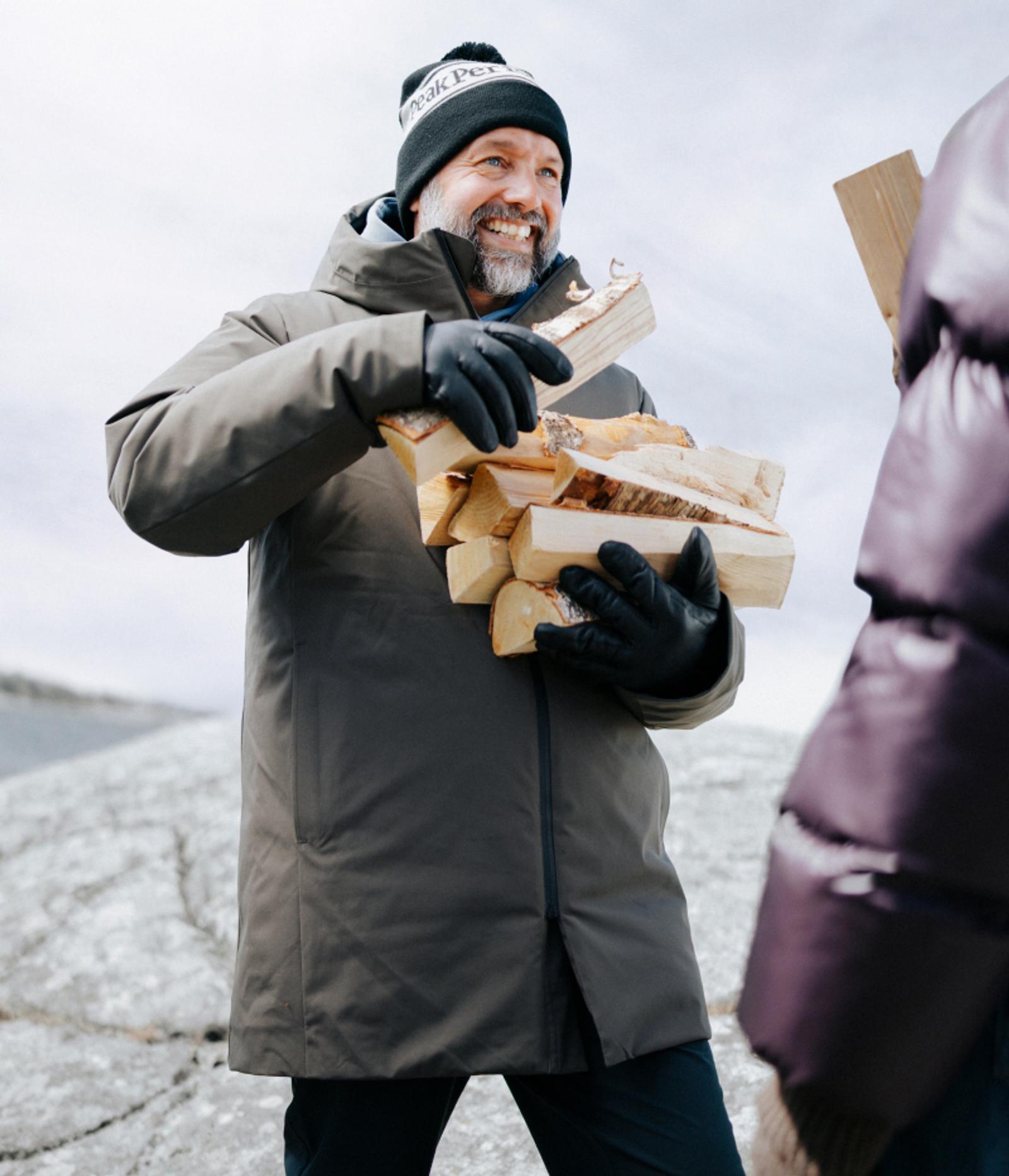 A smiling man in a winter coat and beanie carries a stack of firewood outdoors.