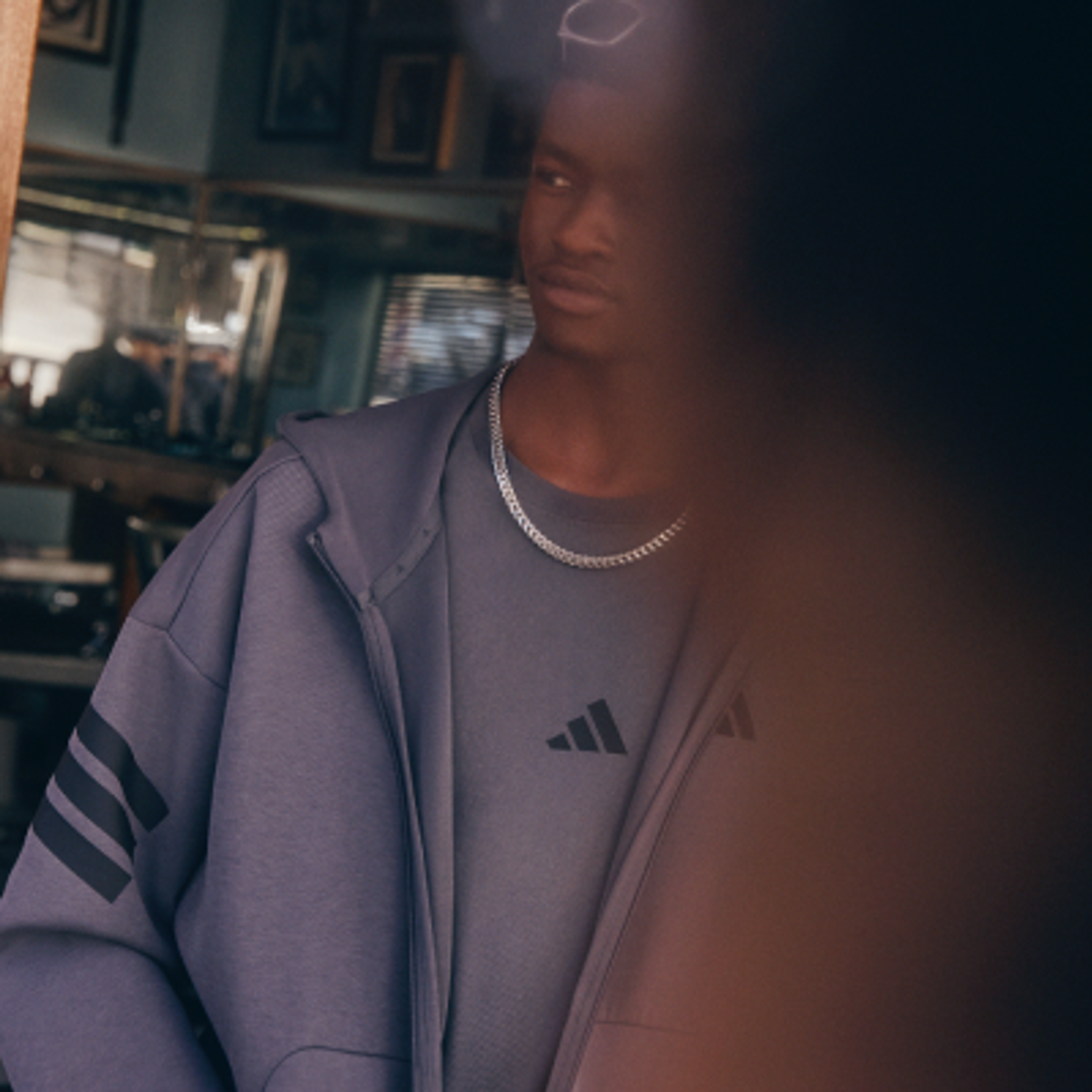 A man in a grey Adidas zip-up hoodie with black stripes on the sleeve, an Adidas logo t-shirt, and a silver chain.