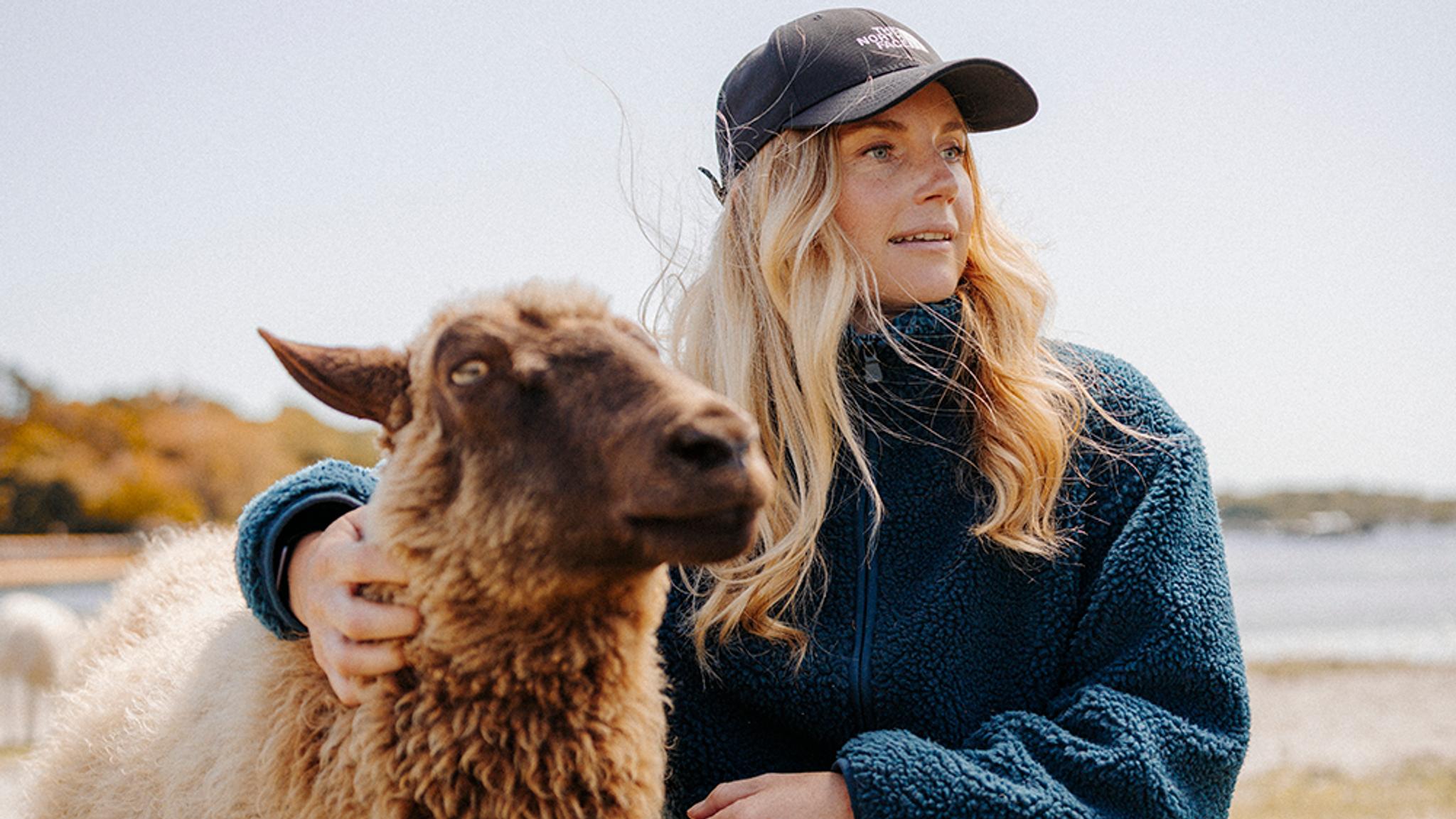 A woman in a cap and fleece jacket pets a sheep outdoors.