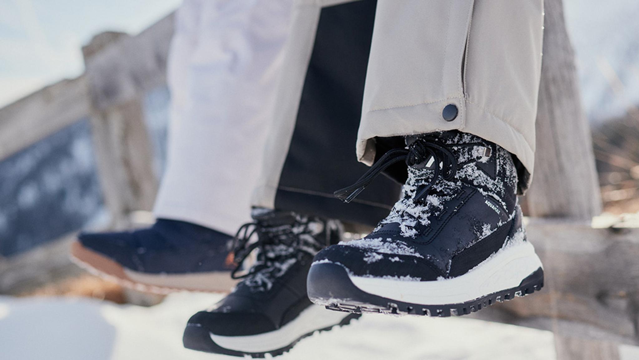 Two pairs of feet dangling above snow; one wears a black, snow-covered winter boot, the other a navy casual shoe.