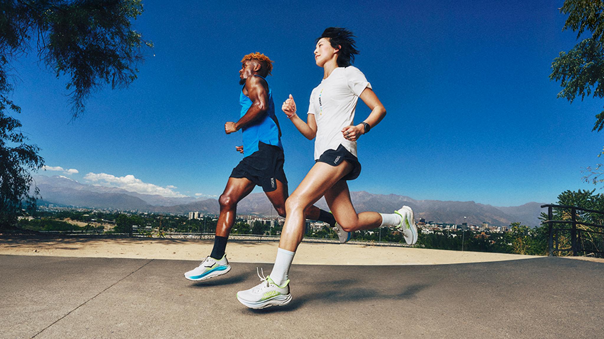 Two runners, a man and a woman, run on a sunny day with mountains and a city in the background.