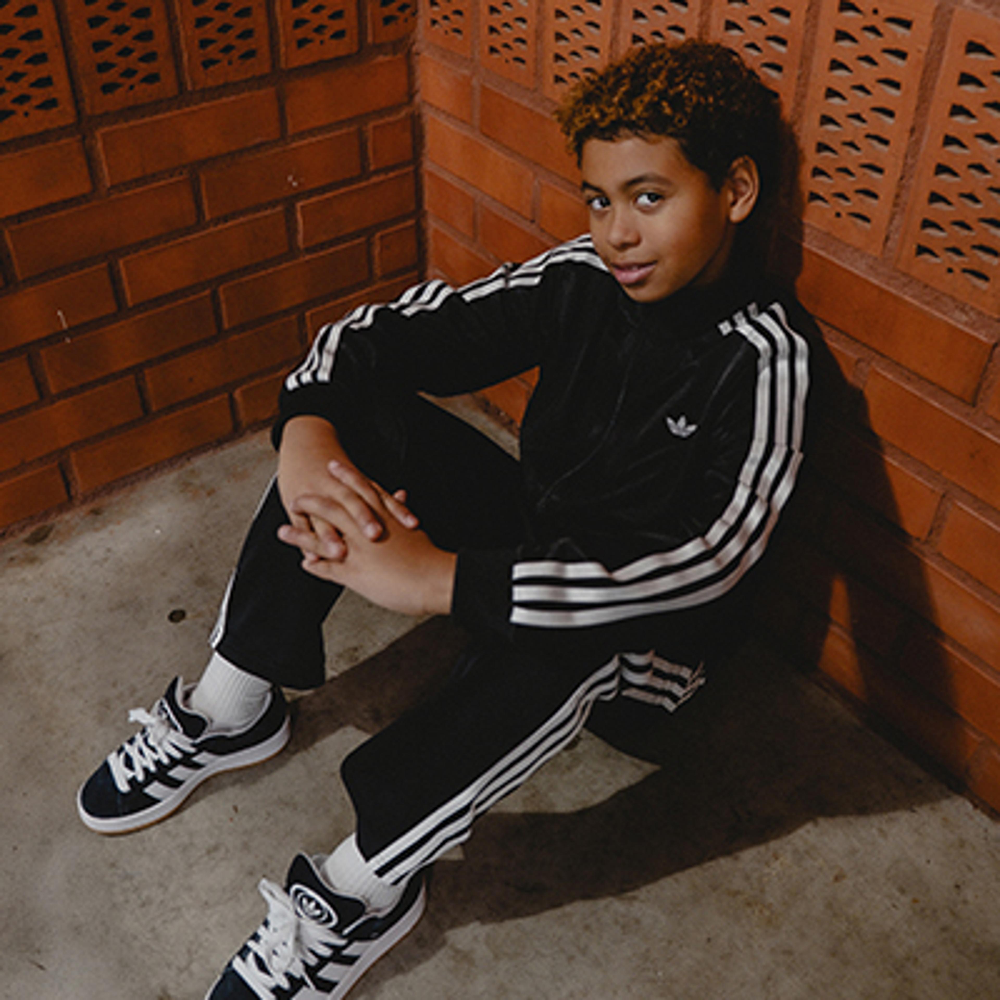 A young person in a black Adidas tracksuit and sneakers sits on a concrete floor against a red brick wall.