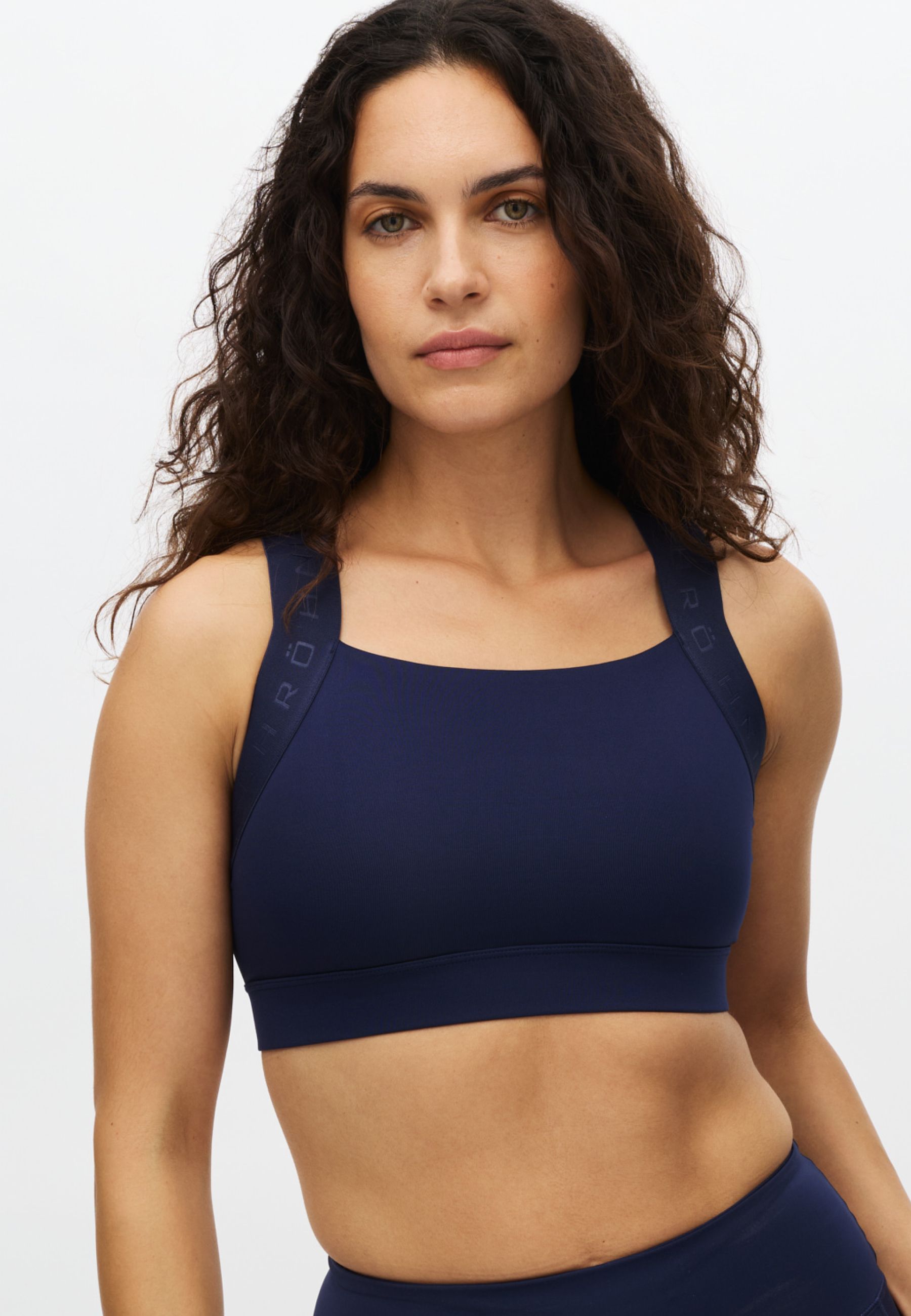 A woman with dark curly hair wears a navy blue sports bra with RÖT branding on the straps.