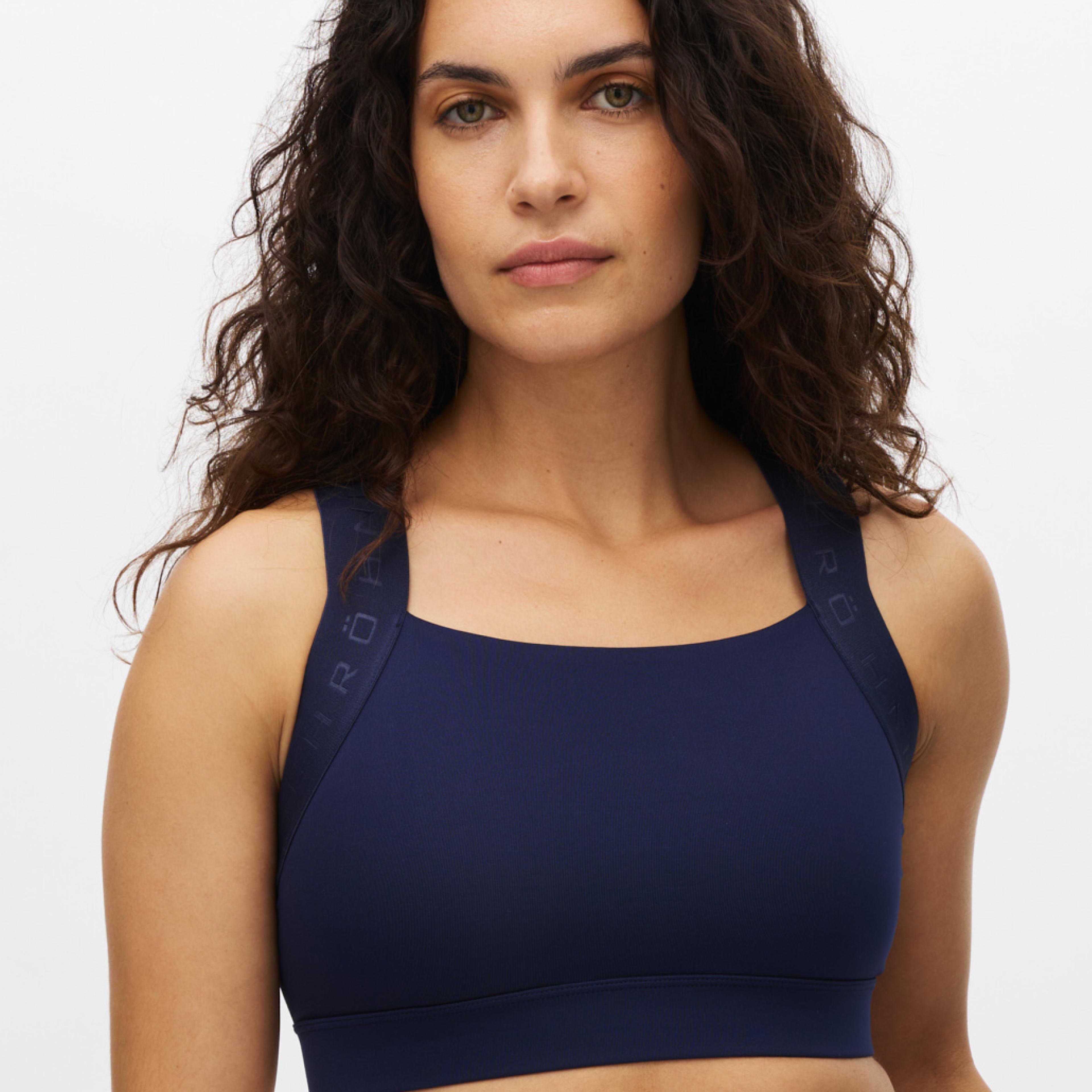 A woman with dark curly hair wears a navy blue sports bra with RÖT branding on the straps.