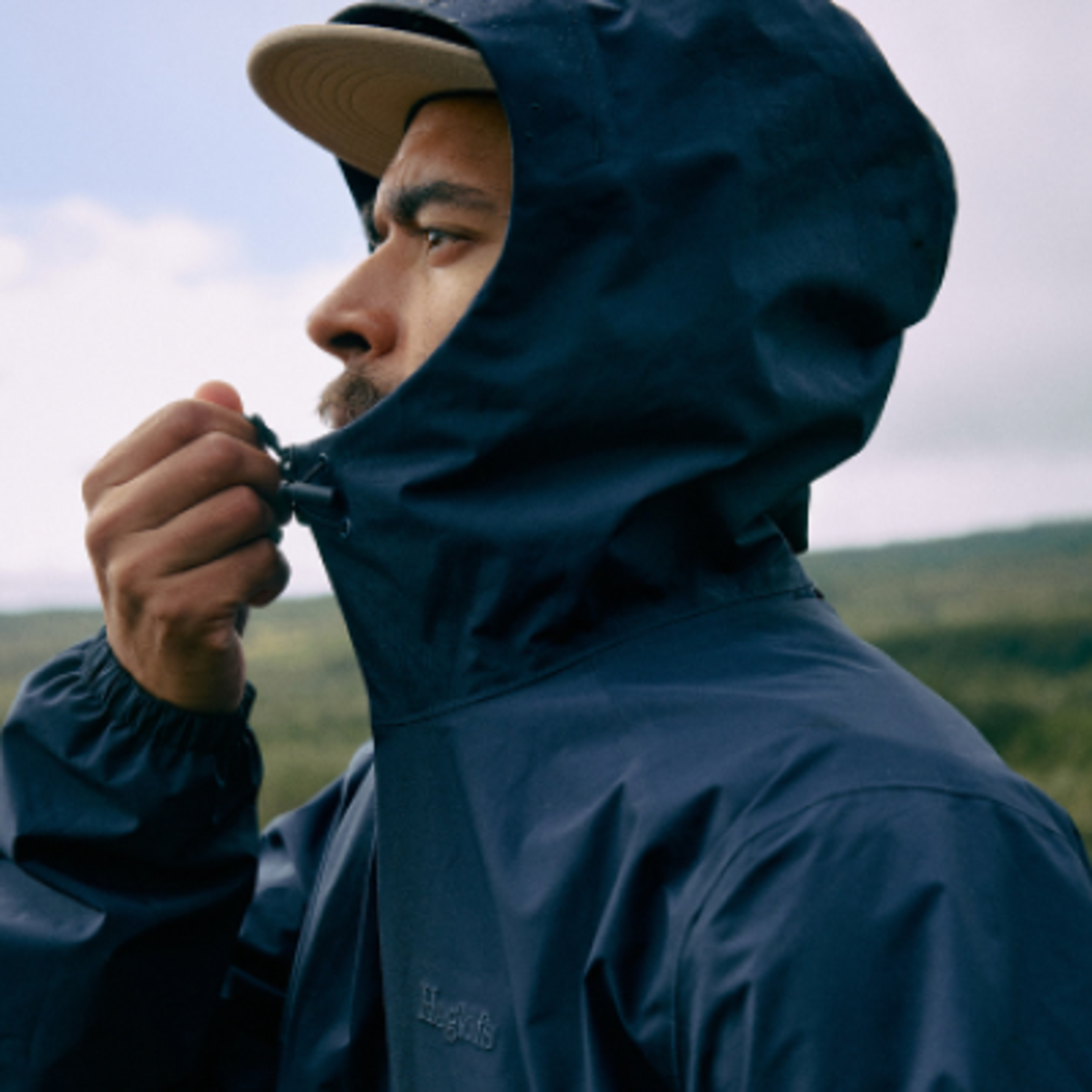 A man in a Haglofs navy hooded jacket and cap adjusts the hood outdoors.