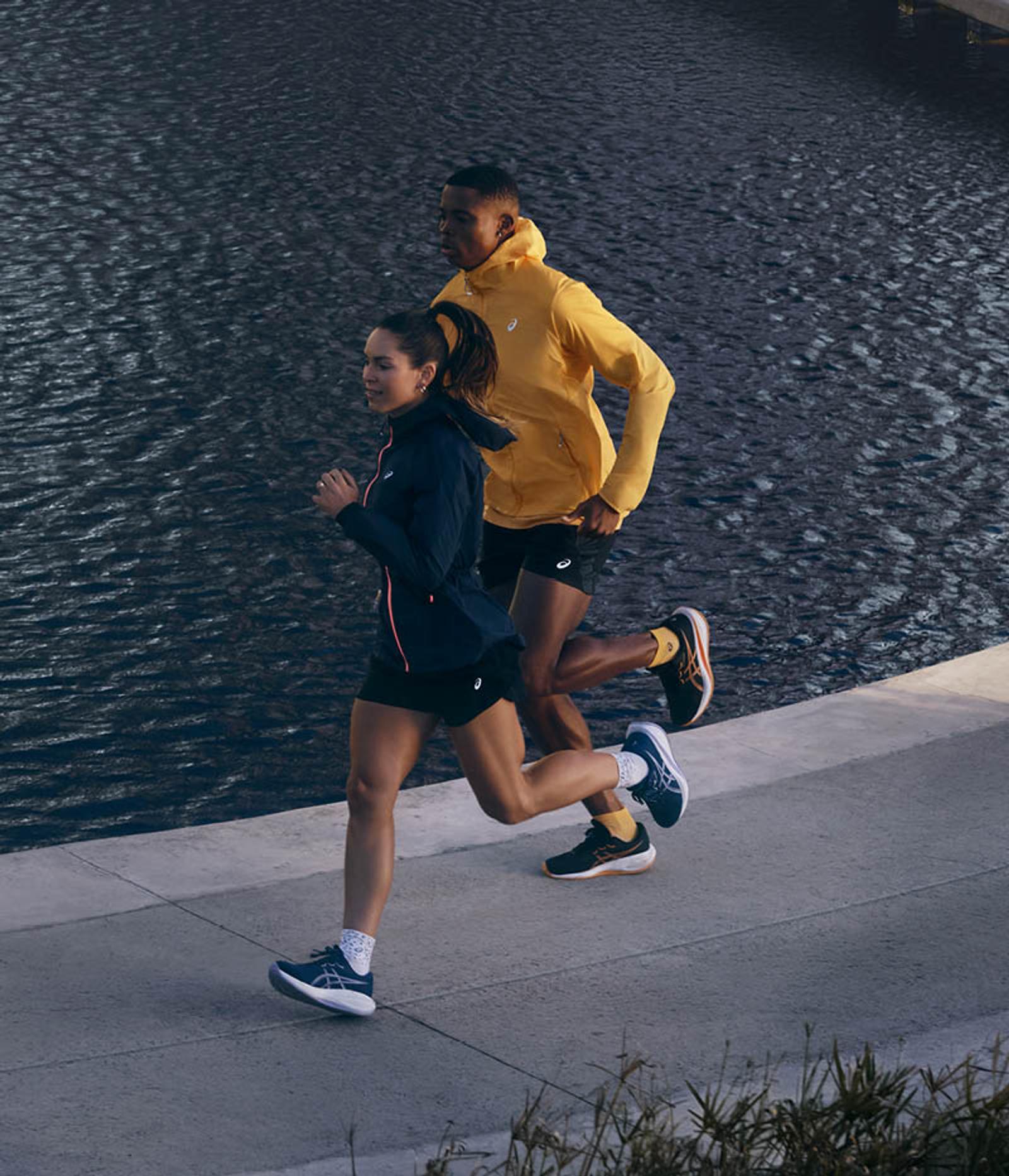 A woman in a dark blue jacket and a man in a yellow jacket run on a paved path next to dark water.