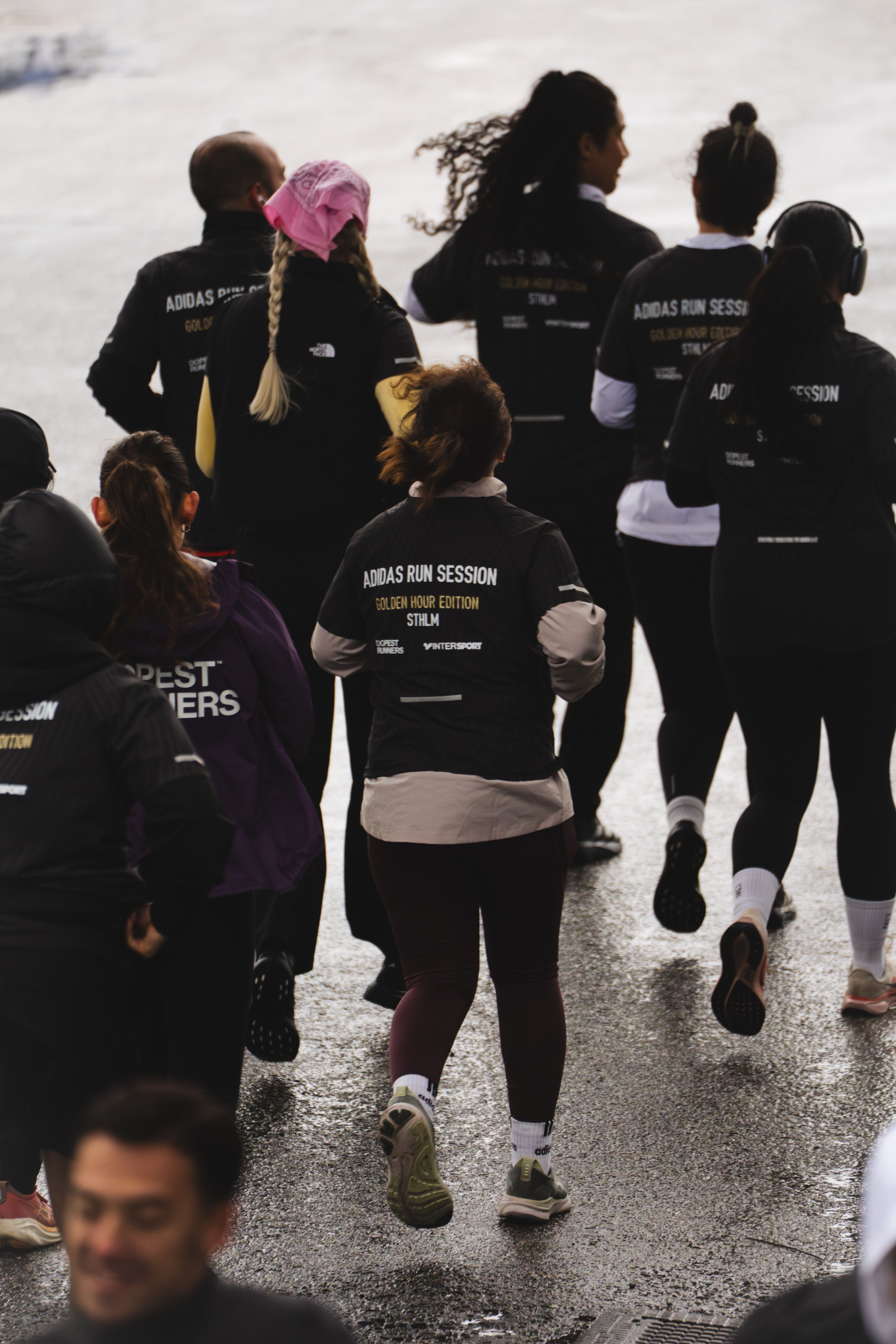 A group of people run on a wet paved surface, many wearing black shirts branded "Adidas Run Session."