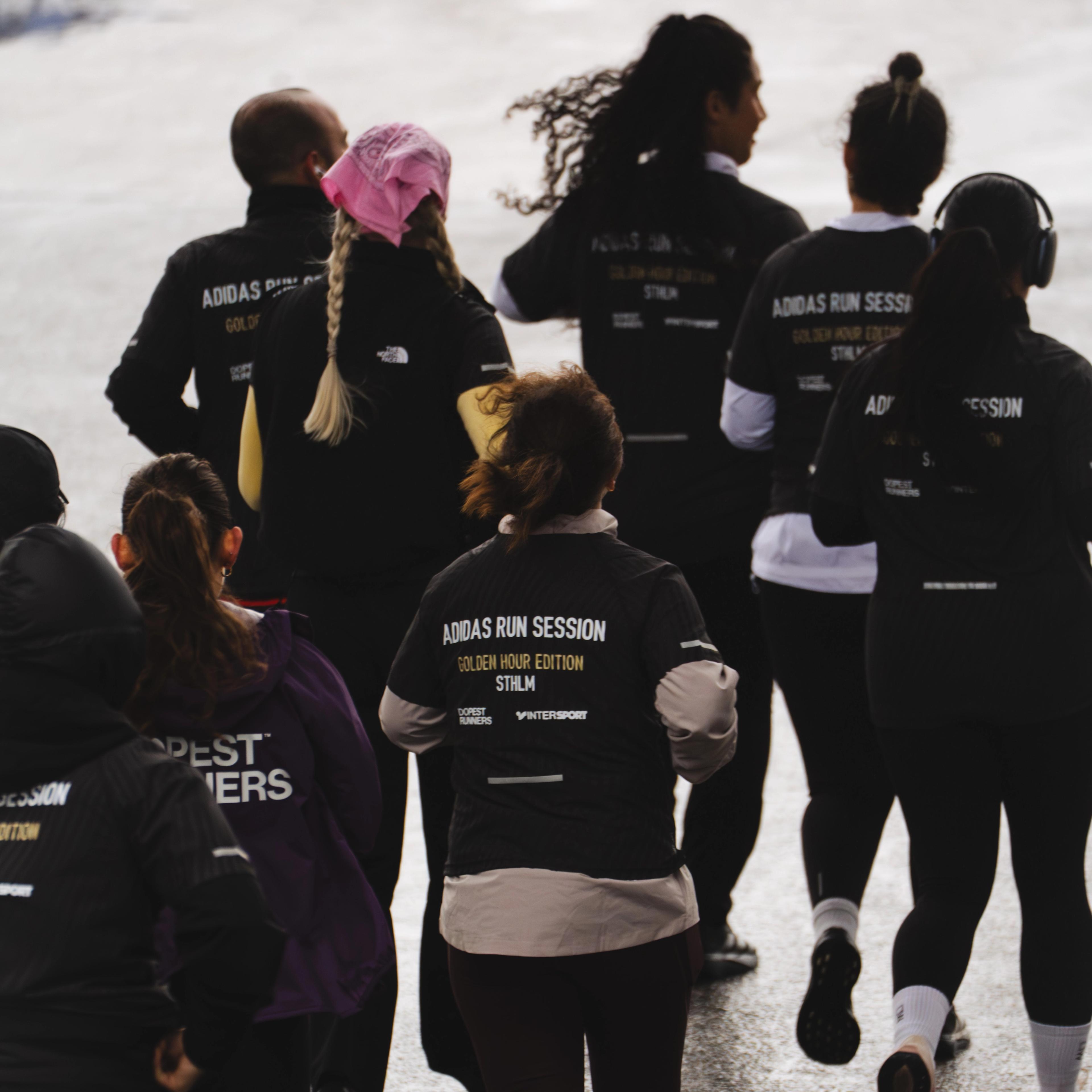 A group of people run on a wet paved surface, many wearing black shirts branded "Adidas Run Session."