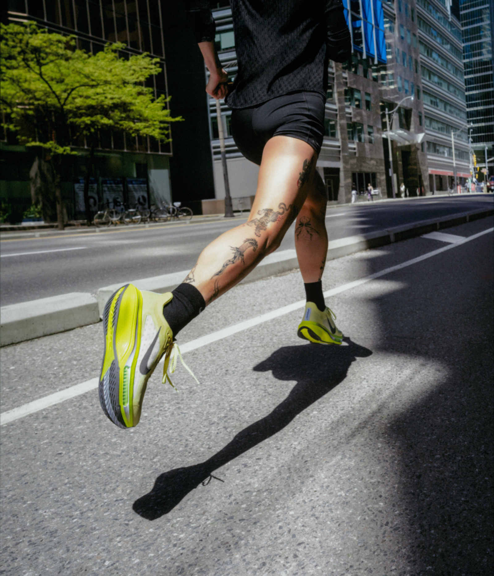A person's tattooed legs running on an urban street in bright yellow running shoes, casting a long shadow.
