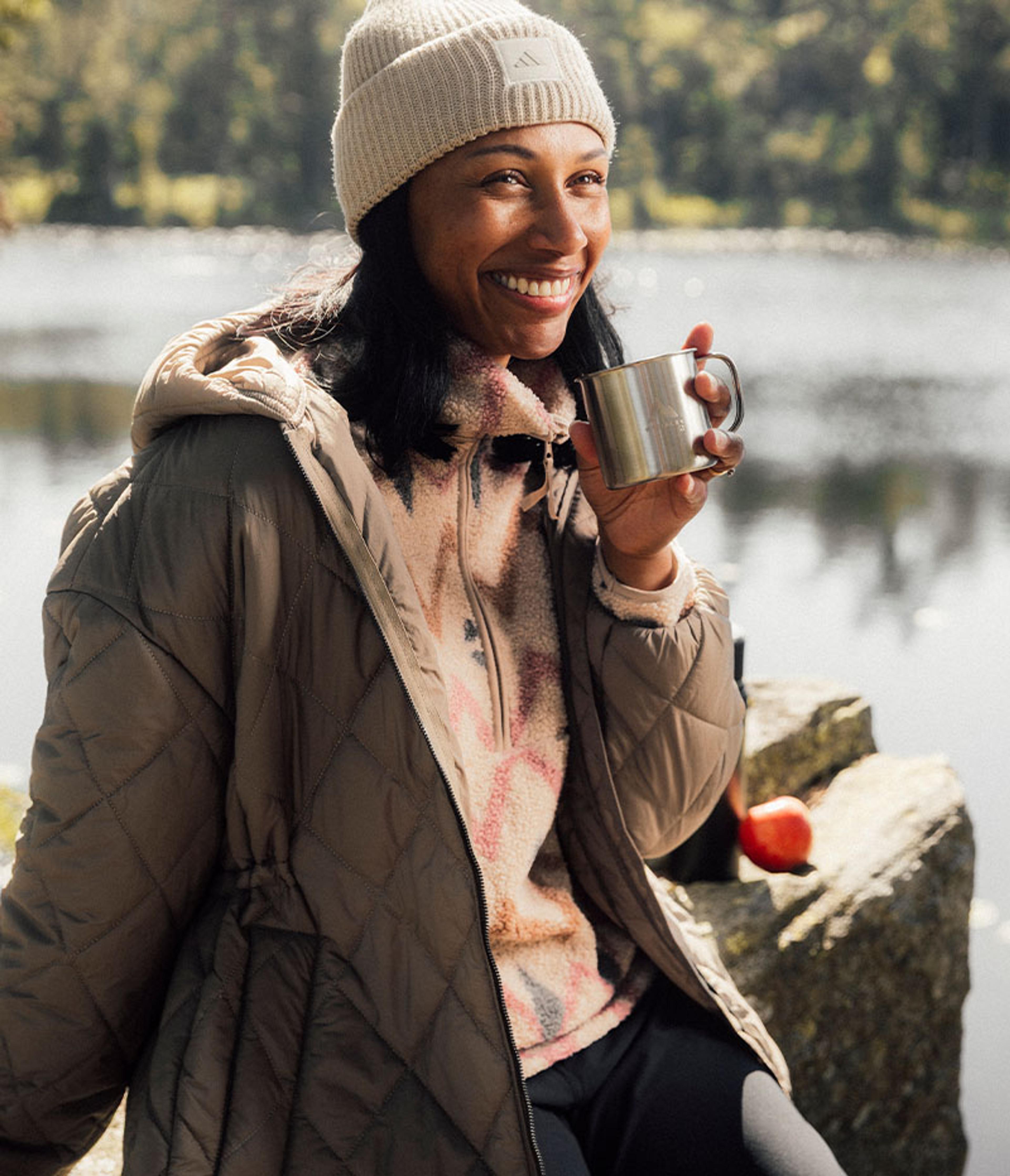a woman is sitting on a rock near a lake holding a cup of coffee .