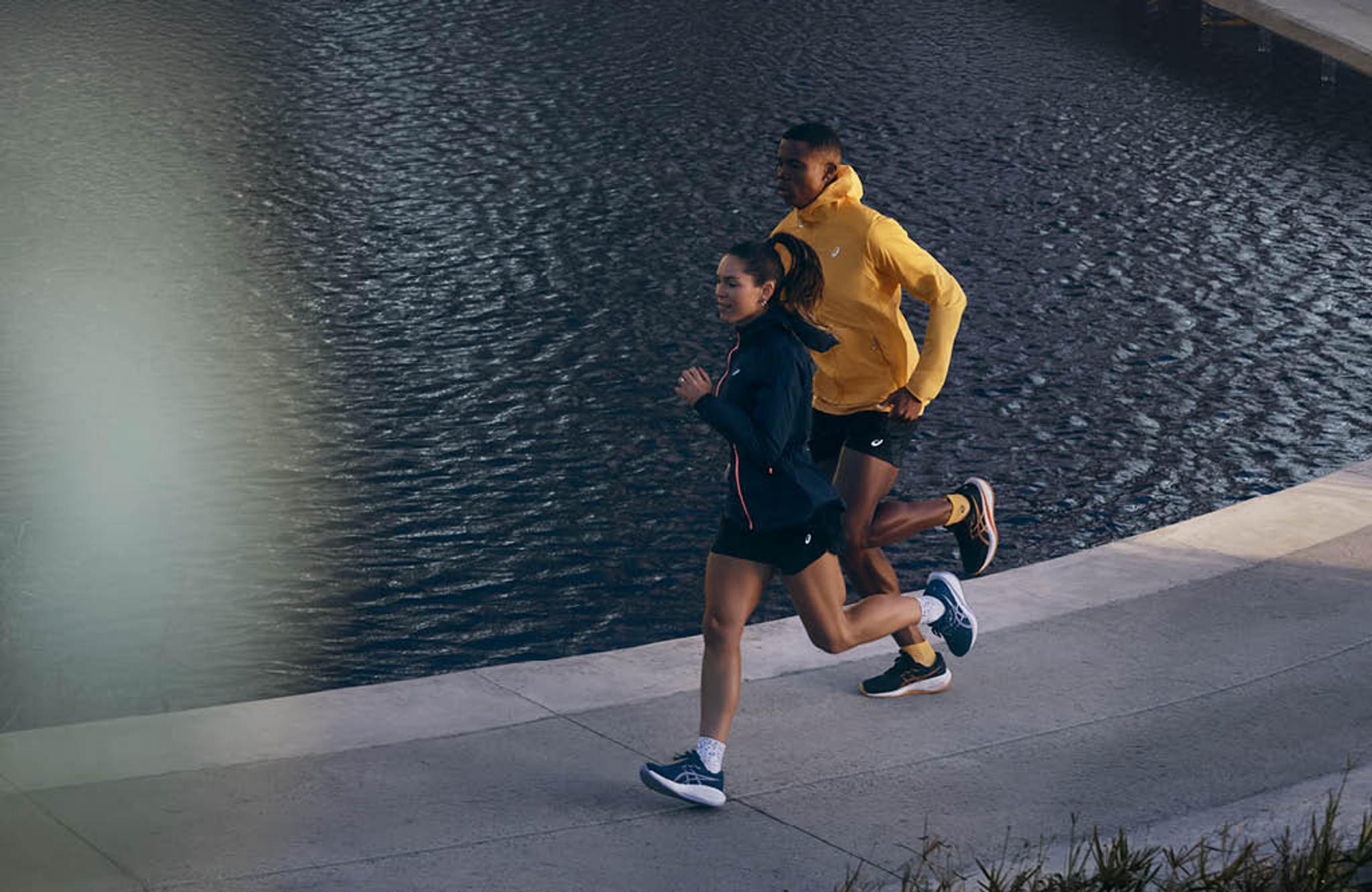 A woman in a dark jacket and a man in a yellow jacket running along a path next to water.