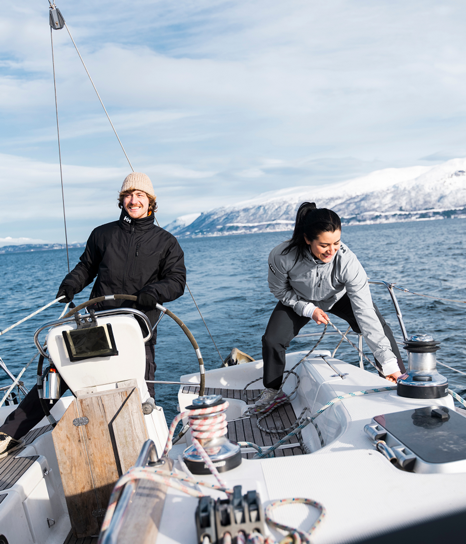 A man steers a sailboat while a woman adjusts ropes, both smiling, with snow-capped mountains in the background.